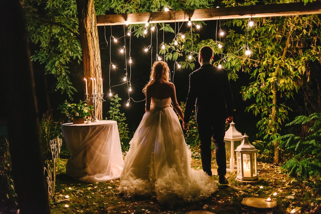 Night wedding ceremony with candles, lanterns and lamps on tree. Bride and groom holding hands on background of baulb lights, back view. Beautiful young couple standing under a tree at night